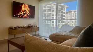 Sala de estar com televisão de tela plana exibindo uma lareira, sofá, mesa de centro e uma janela grande com vista para edifícios de apartamentos.