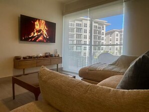 A living room with a flat-screen TV displaying a fire, a sofa, a coffee table, and a large window with a view of apartment buildings. - Apartamento frente al mar con vista a la piscina (punta chame)