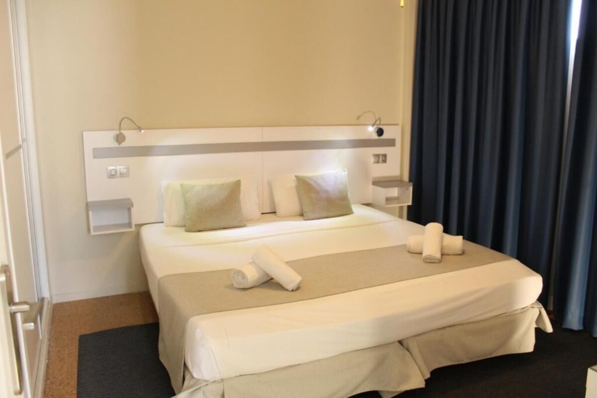 A hotel room with a large bed, a headboard with a light fixture, a bedside table, and a window with curtains.