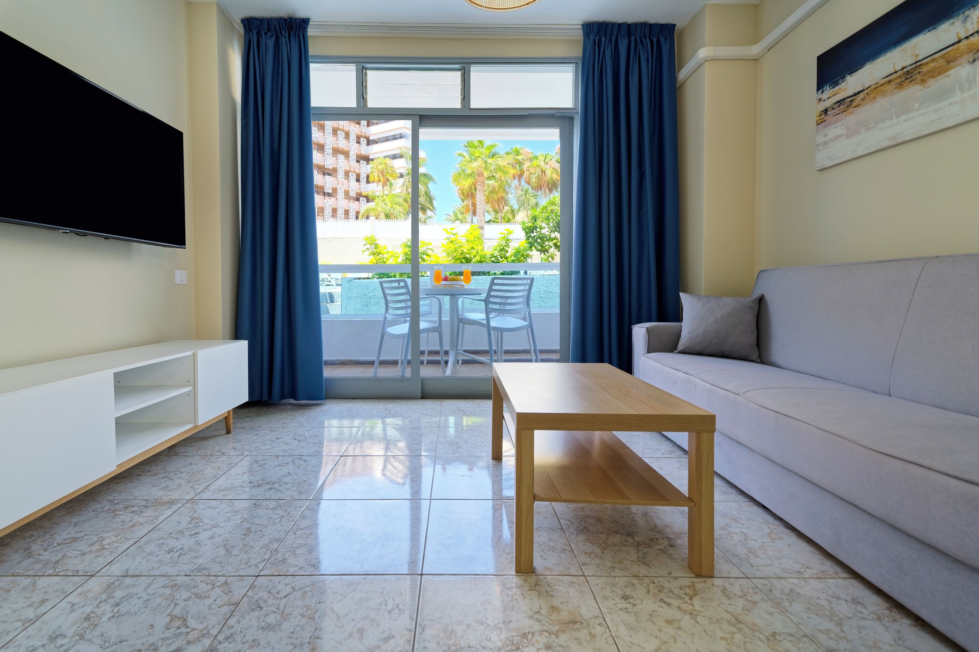 Uma sala de estar moderna com televisão de tela plana, mesa de centro de madeira, sofá cinza e uma varanda com cortinas azuis.