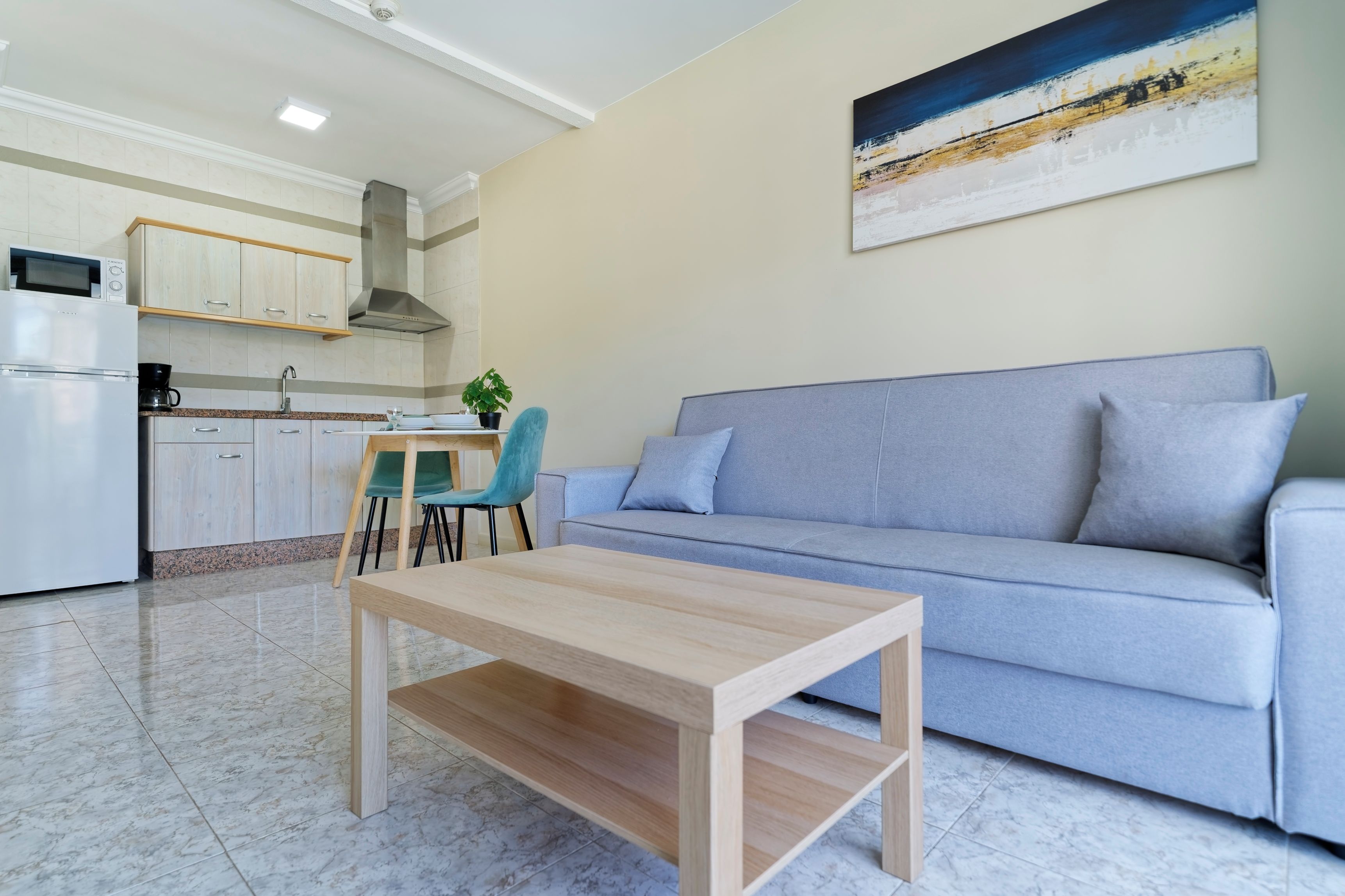 A modern living room with a grey sofa, a wooden coffee table, and a kitchen area with cabinets and a microwave.