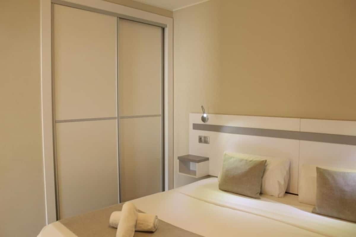 A hotel room with a bed, pillows, a nightstand, a wall-mounted light, and a sliding door.