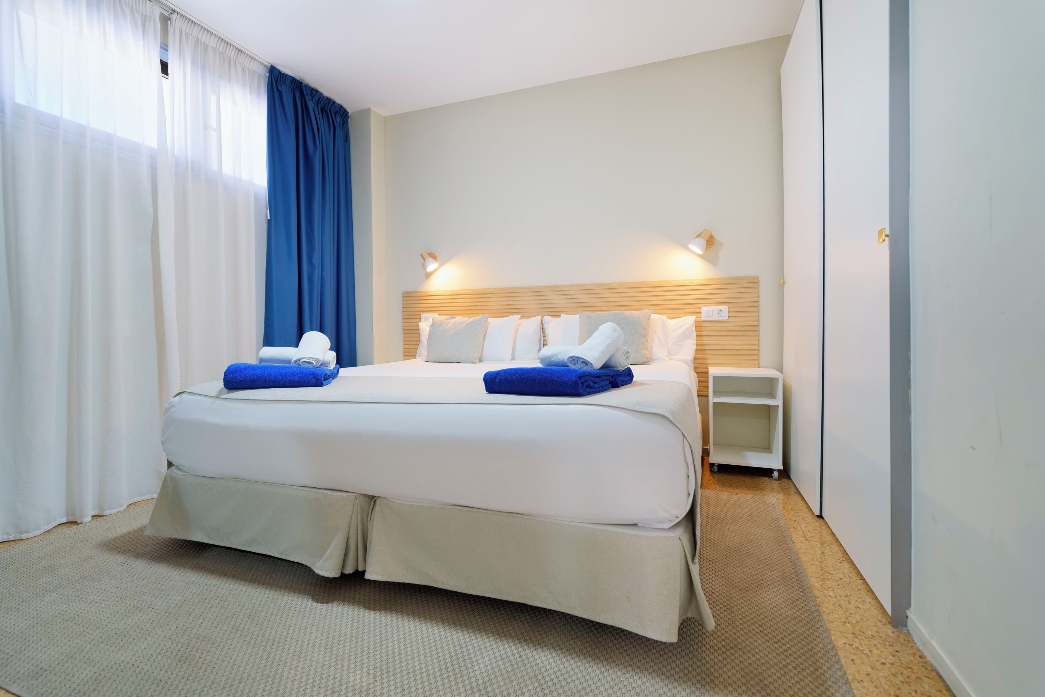 A hotel room with a large bed, two bedside tables, a wall-mounted lamp, and a window with curtains.