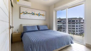 Quarto com uma cama grande, mesa de cabeceira, pintura e vista de um prédio através de uma janela ampla.