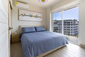 Ein Schlafzimmer mit einem großen Bett, einem Nachttisch, einem Gemälde und Blick auf ein Gebäude durch ein großes Fenster.