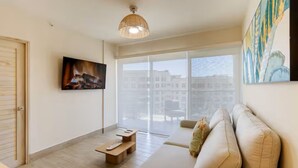 Uma sala de estar moderna com sofá, mesa de centro, TV fixada na parede e uma grande janela com vista para edifícios.