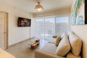 Ein modernes Wohnzimmer mit einer Couch, einem Couchtisch, einem an der Wand montierten Fernseher und einem großen Fenster mit Blick auf Gebäude.