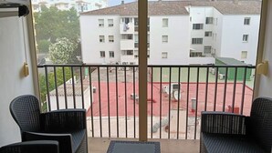 Un balcón con dos sillones de mimbre y una mesita, con vistas a un patio que incluye edificios y una zona de juegos roja.