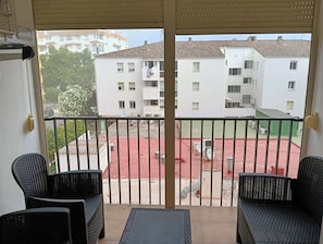 Ein Balkon mit zwei Korbstühlen und einem kleinen Tisch, mit Blick auf einen Innenhof mit Gebäuden und einem roten Spielbereich.