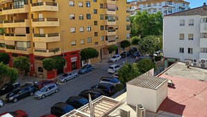 Un edificio amarillo de varios pisos con balcones, un estacionamiento con distintos vehículos y un edificio blanco al fondo.