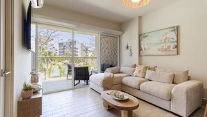 Una sala de estar con sofá, mesa de centro, televisor y balcón con vista a edificios y árboles.