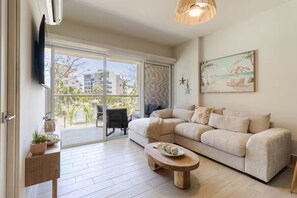 Un salon comprenant un canapé, une table basse, un téléviseur et un balcon offrant une vue sur les immeubles et les arbres.