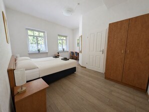 Ein Schlafzimmer mit Bett, Nachttisch, Lampe, zwei Fenstern, einem Kleiderschrank und einer Tür.