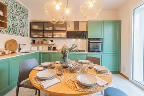 Zona pranzo con tavolo rotondo apparecchiato per quattro persone, pensili verdi, cucina sullo sfondo e lampade a sospensione.