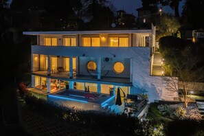 Una moderna casa a due piani con un balcone, una piscina e un esterno ben illuminato.