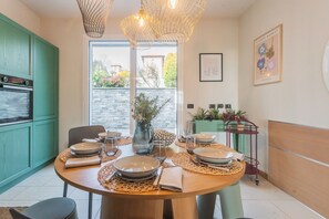 A dining area with a round table set for four, a kitchen with green cabinets, and a view through a glass door. - Villa Olivo with garden & pool (Montano Lucino)