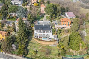 A modern two-story house with a large balcony, surrounded by trees and a small garden.
