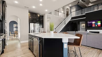 A modern kitchen with a central island, a dining area with chairs, a staircase, and a living area with a couch and a TV.