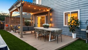 Een overdekt terras met een tafel en stoelen, een pergola met sfeerverlichting en een huis met een raam.