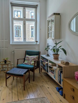 Ein Zimmer mit Holzboden, einem Sessel mit Kissen, einem Bücherregal, einem Heizkörper und einem Fenster mit Blick auf Gebäude.