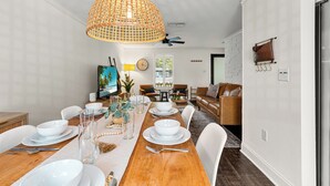 A dining area with a wooden table set for four, white chairs, a large woven pendant light, and a television in the background. - Mins to Beaches/Downtown, Quiet Neighborhood, Community Pool (Sarasota)