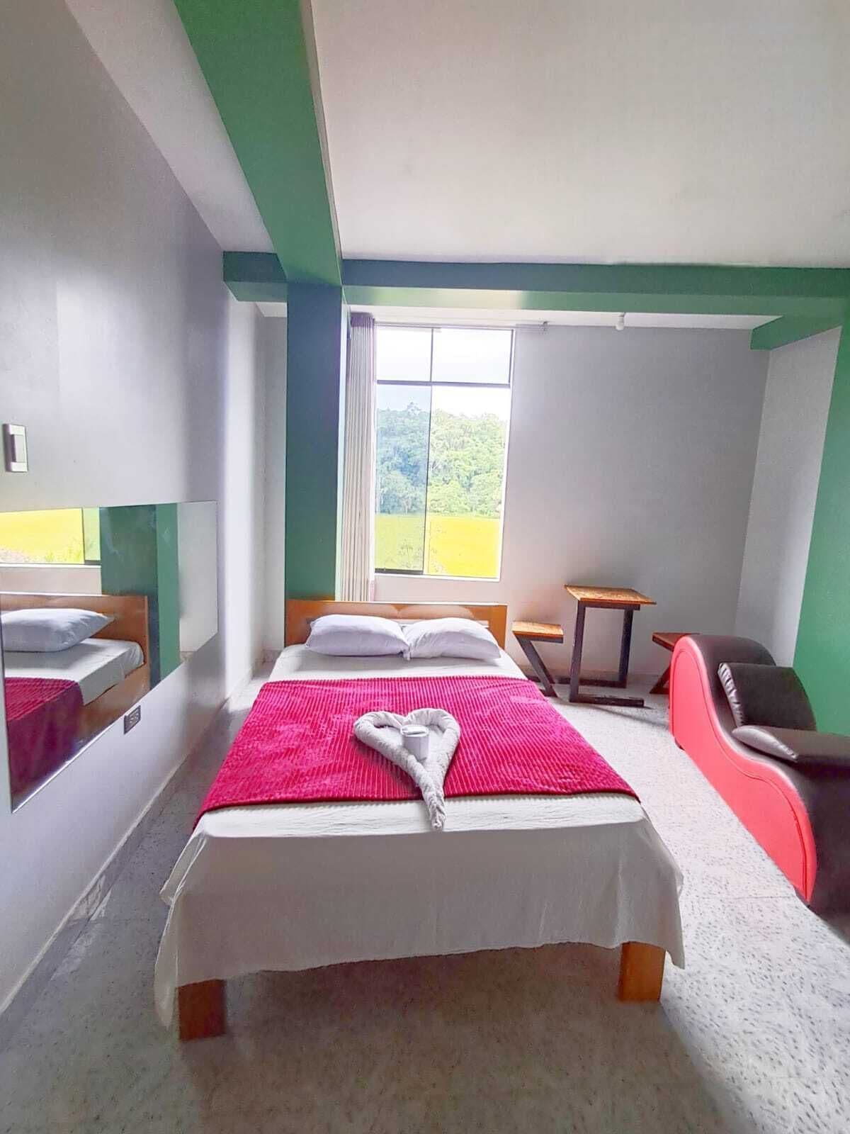 Hotellihuone, jossa on sänky, tuoli, pieni pöytä ja ikkuna, josta avautuu näkymä vehreään maisemaan.
