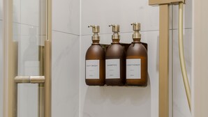 Ein Badezimmer mit einer Duschkabine aus Glas, einem goldfarbenen Duschkopf und drei braunen Toilettenartikeln auf einem an der Wand befestigten Regal.