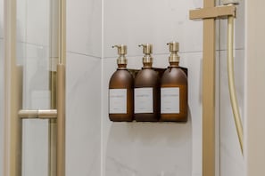 Ein Badezimmer mit einer Duschkabine aus Glas, einem goldfarbenen Duschkopf und drei braunen Toilettenartikeln auf einem an der Wand befestigten Regal.