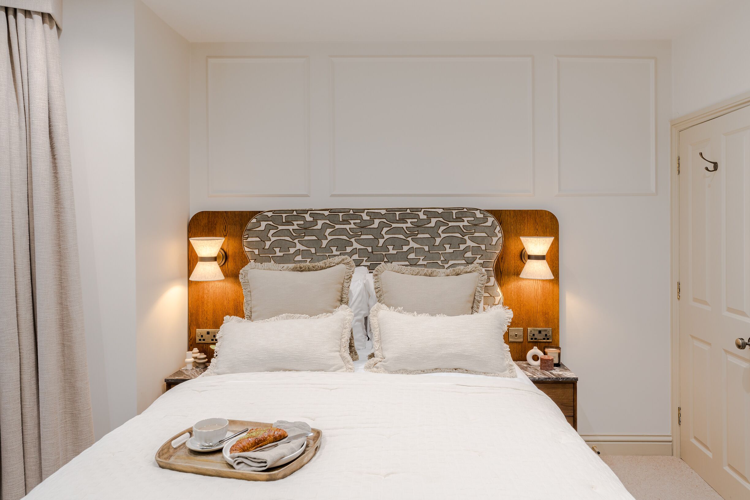 A neatly made bed with a tray of breakfast items, a wooden headboard with patterned cushions, and bedside lamps.