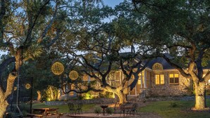 Una casa de piedra con un gran árbol en primer plano, un columpio y una mesa de picnic.