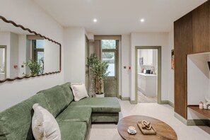Ein modernes Wohnzimmer mit einem grünen Sofa, einem Holztisch, einer Topfpflanze und Blick in die Küche durch eine offene Tür.