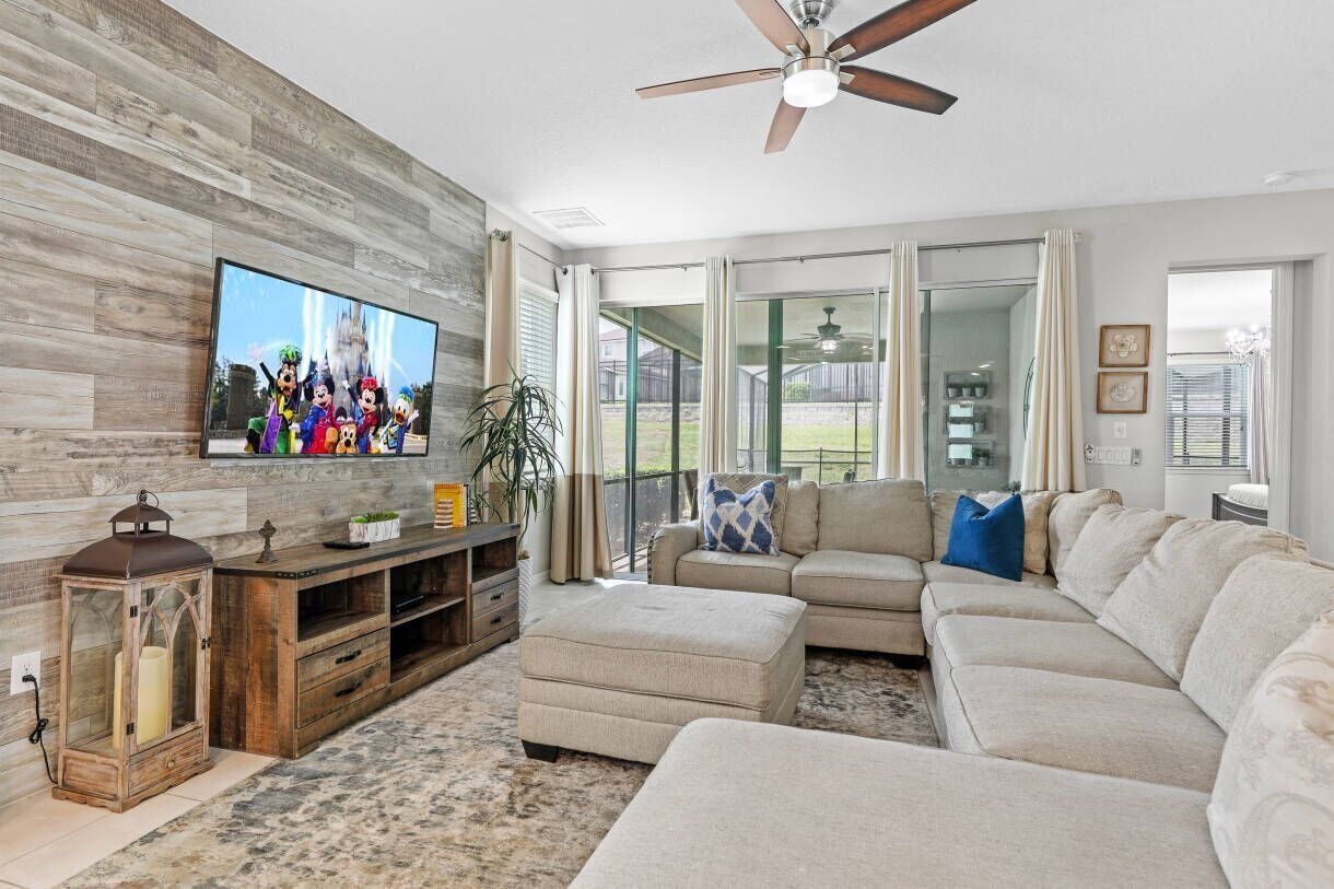 A living room with a large flat-screen TV, a ceiling fan, a wooden entertainment unit, a beige sectional sofa, and a large window with curtains.