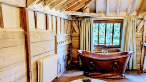 A rustic wooden room with a large bathtub, a radiator, and a window with curtains. - 8 Henham Barns, Henham Park, Nr Southwold, Suffolk (Suffolk)