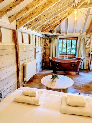 A rustic wooden room with a large bathtub, a radiator, and a window with curtains. - 8 Henham Barns, Henham Park, Nr Southwold, Suffolk (Suffolk)