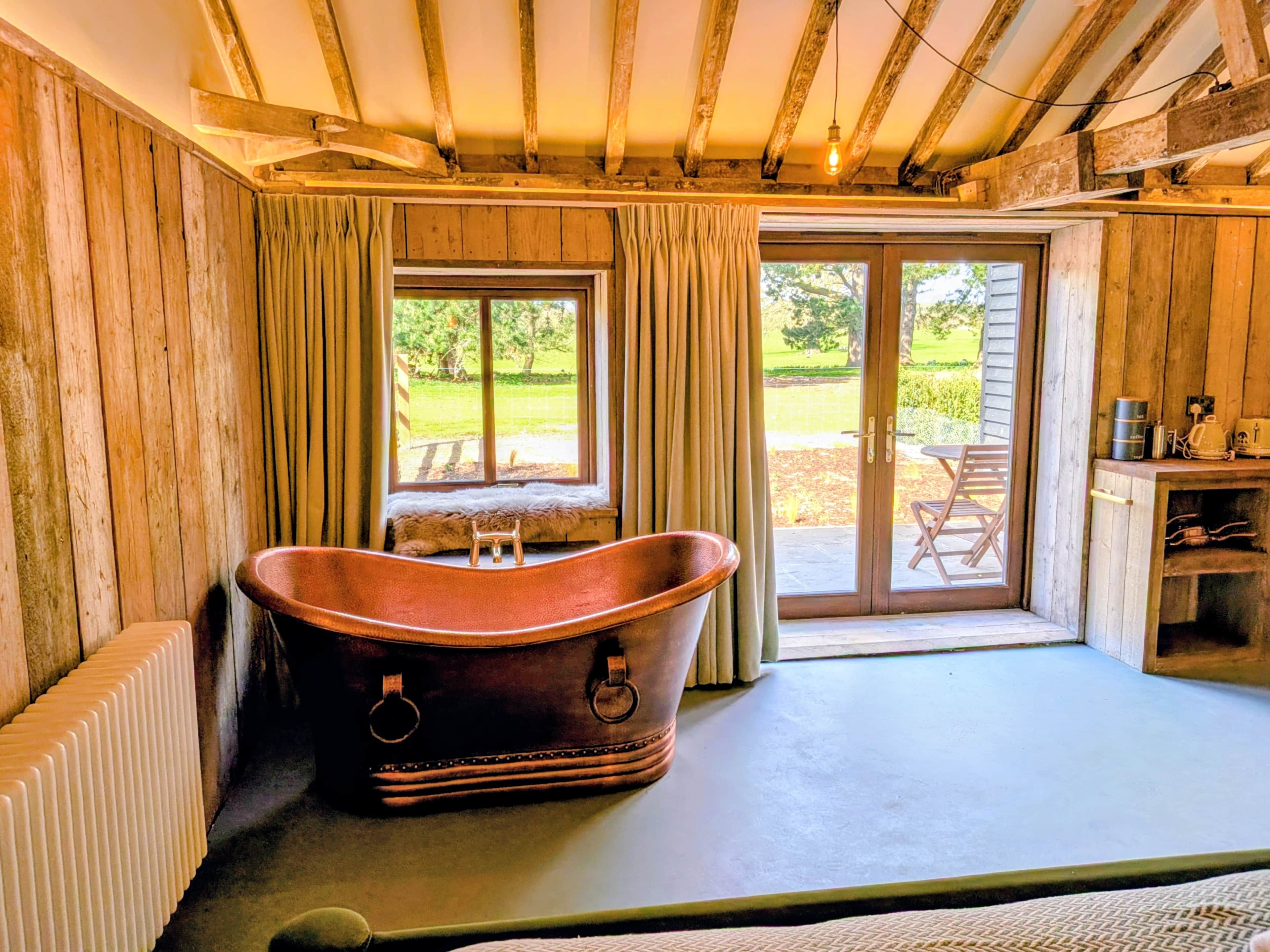 Ein rustikaler Raum mit einer großen Badewanne, Holzwänden und Blick auf einen Garten durch eine Schiebetür.