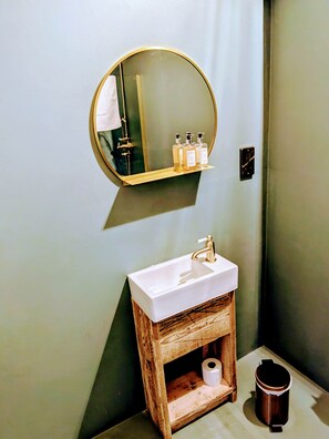 Et badeværelse med en hvid vask, en trækommode, et rundt spejl og toiletartikler på en hylde.