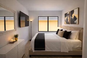 Une chambre à coucher comprenant un lit, une table de chevet, une lampe, une fenêtre et une œuvre d’art.
