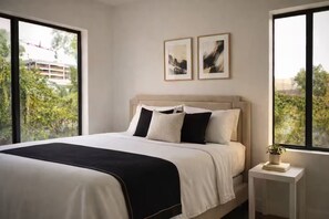 A bedroom with a bed, two framed pictures on the wall, a nightstand with a plant, and large windows with a view of greenery.