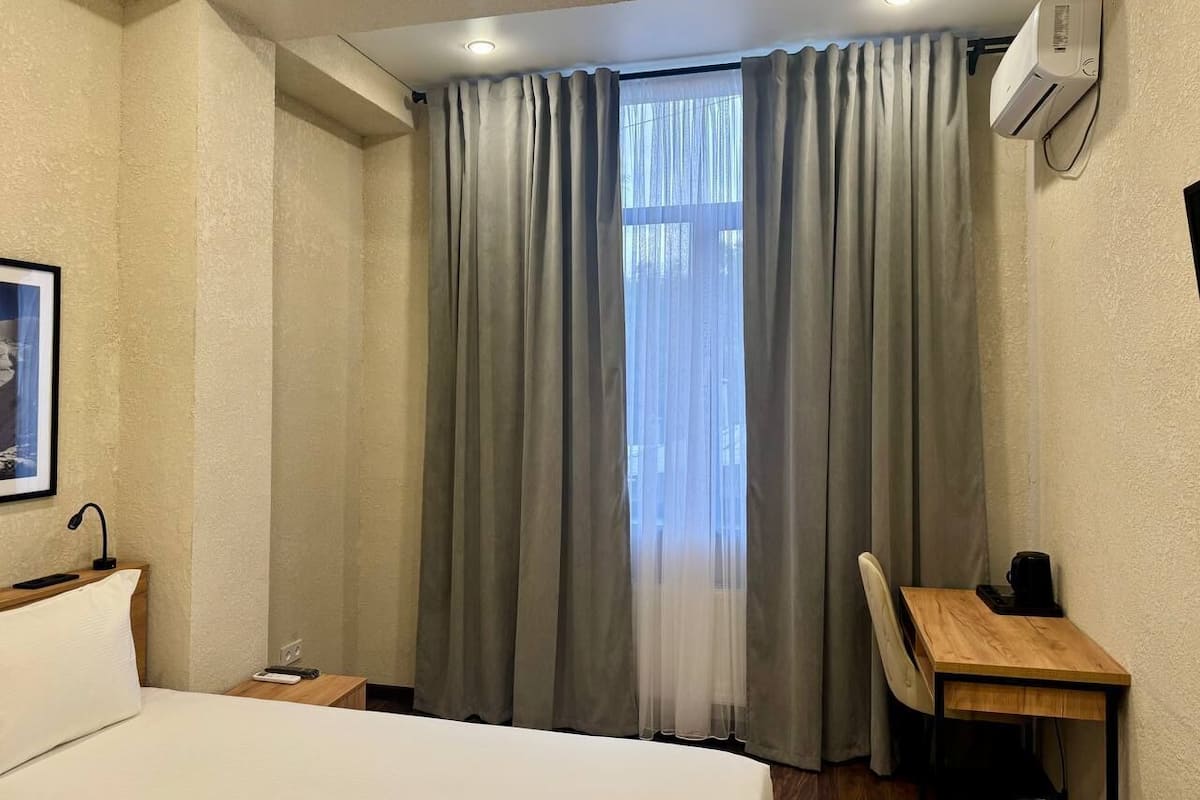 A hotel room with a bed, a desk, a chair, a window with curtains, and an air conditioning unit.