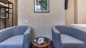 Habitación de hotel con dos sillones grises, una mesita redonda con un control remoto y revistas, y un cuadro en la pared con una imagen de un lago de montaña.