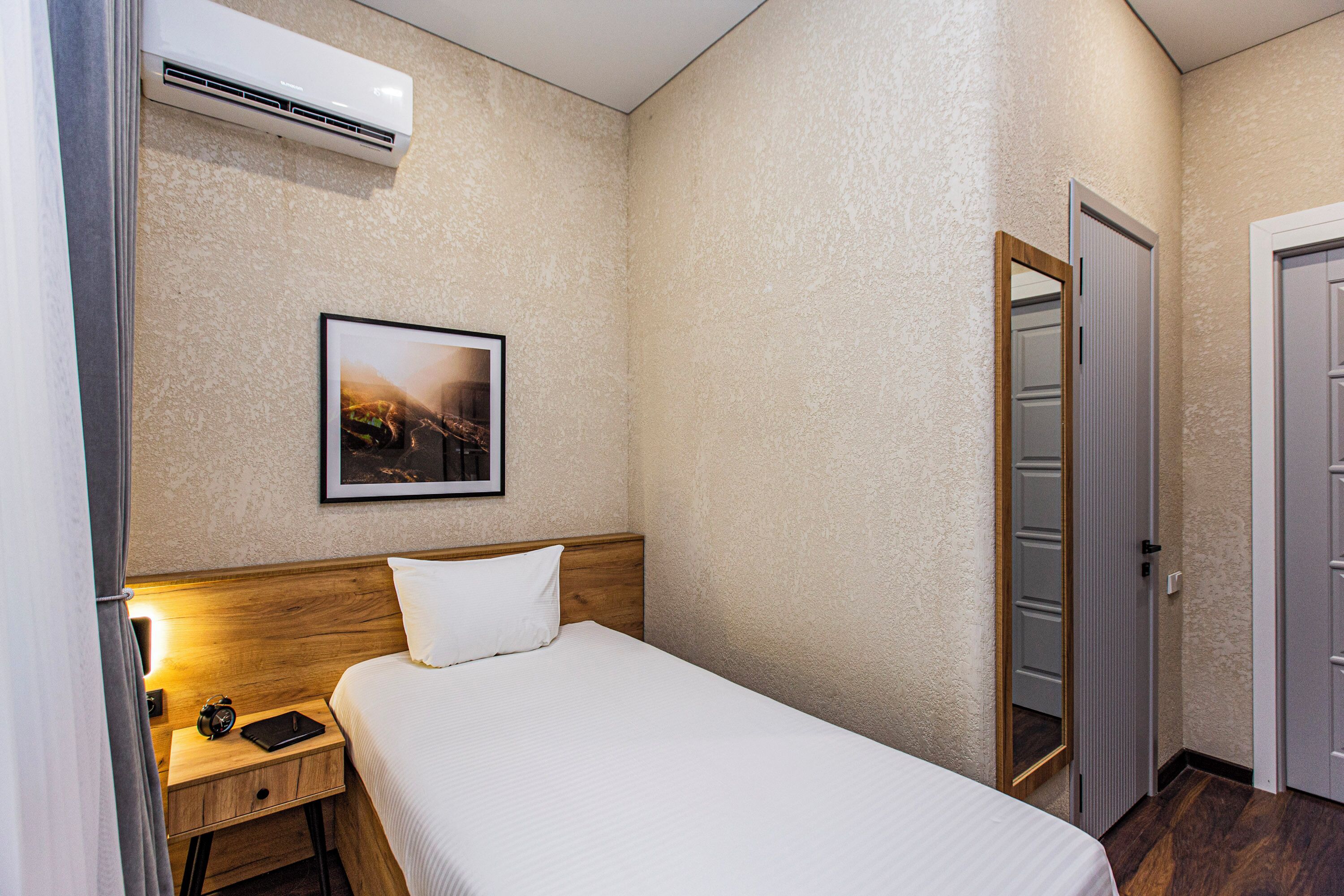 A hotel room with a bed, a nightstand, a framed picture, an air conditioning unit, and a door.