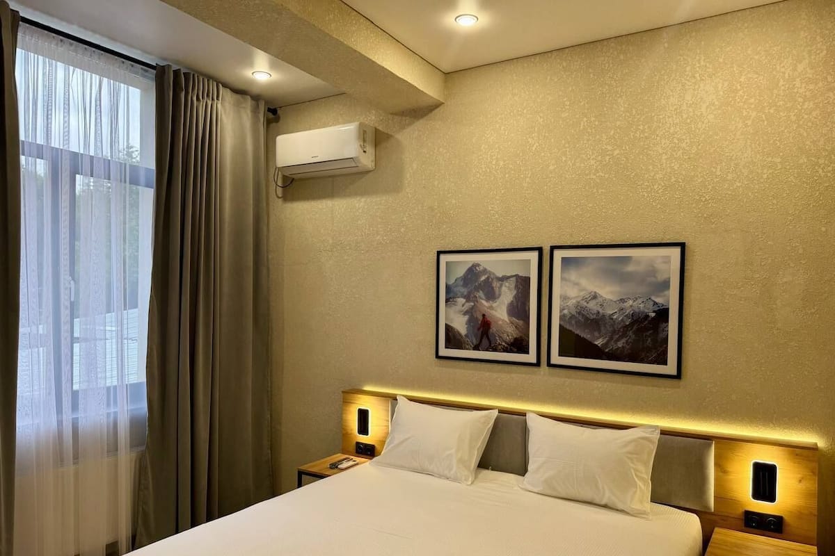 A hotel room with a large bed, two wall-mounted pictures, an air conditioning unit, and a window with curtains.