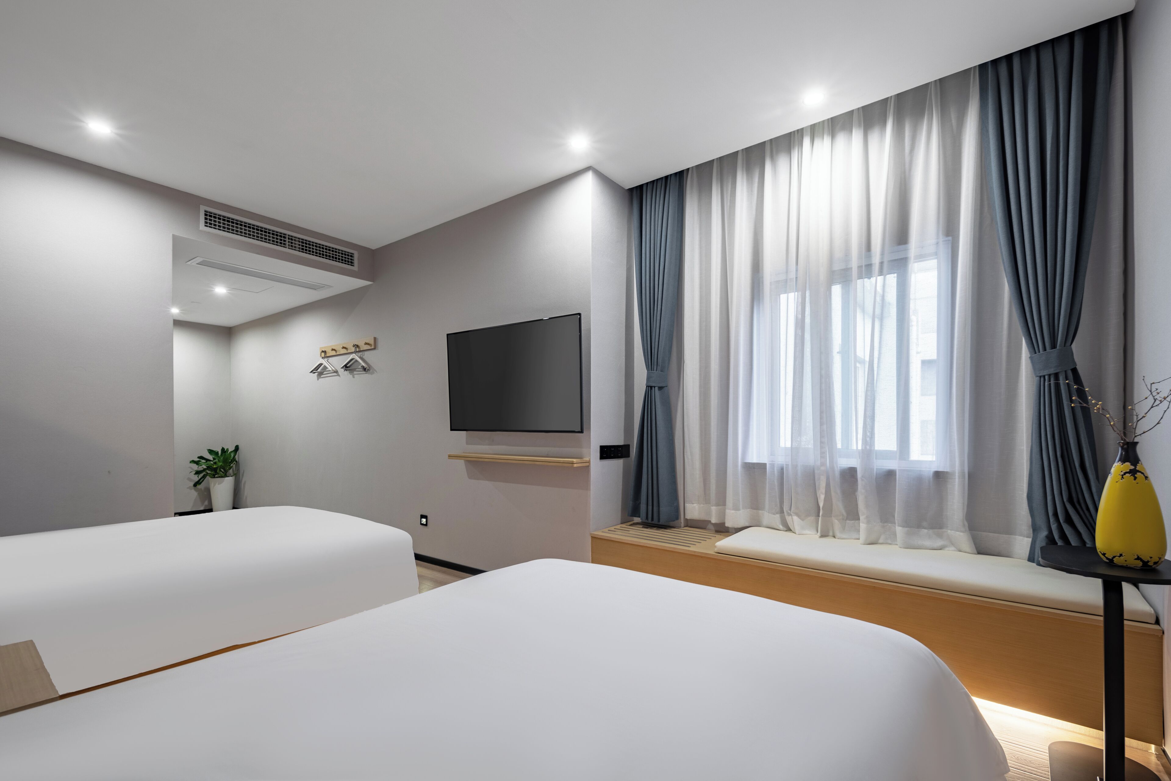 A modern hotel room with two beds, a flat-screen TV, a potted plant, a window with curtains, and a bench.