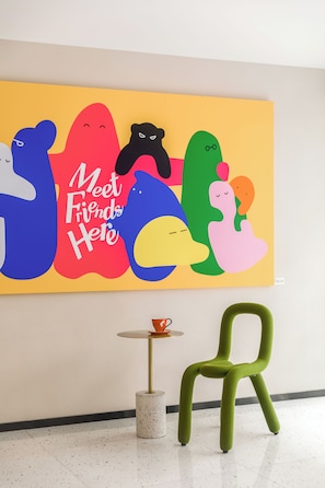 Un cuadro abstracto y colorido con el texto 'Meet Friends Here' en una pared amarilla, una silla verde y una pequeña mesa redonda con una taza.