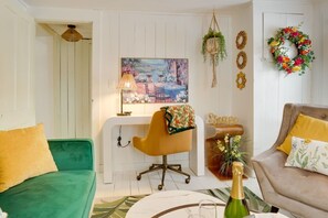 Ein Wohnzimmer mit grünem Sofa, gelbem Sessel, beigem Sofa, einem Gemälde, einem Schreibtisch, einer hängenden Pflanze und einem Kranz an der Wand.