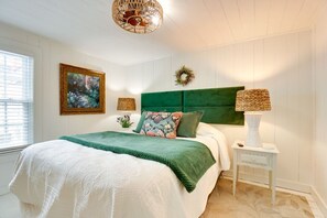Une chambre à coucher avec une tête de lit recouverte d’un tissu vert, un grand lit avec une couette blanche, une table de chevet avec une lampe et un tableau floral encadré accroché au mur.