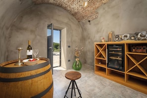 Un interior rústico con un barril de vino, una botella, una copa, un jarrón verde con flores secas y una botellera de madera.