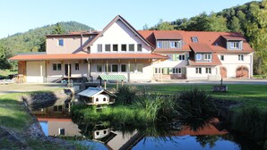Ein großes, zweistöckiges Haus mit Teich davor, umgeben von Grünflächen und Hügeln.