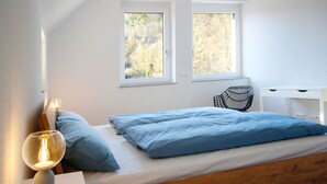 Ein Schlafzimmer mit einem Bett, zwei Fenstern, einer Nachttischlampe, einem Sessel und einer weißen Kommode.