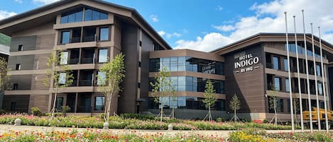 A modern hotel building with multiple floors, balconies, and a landscaped garden with colorful flowers.